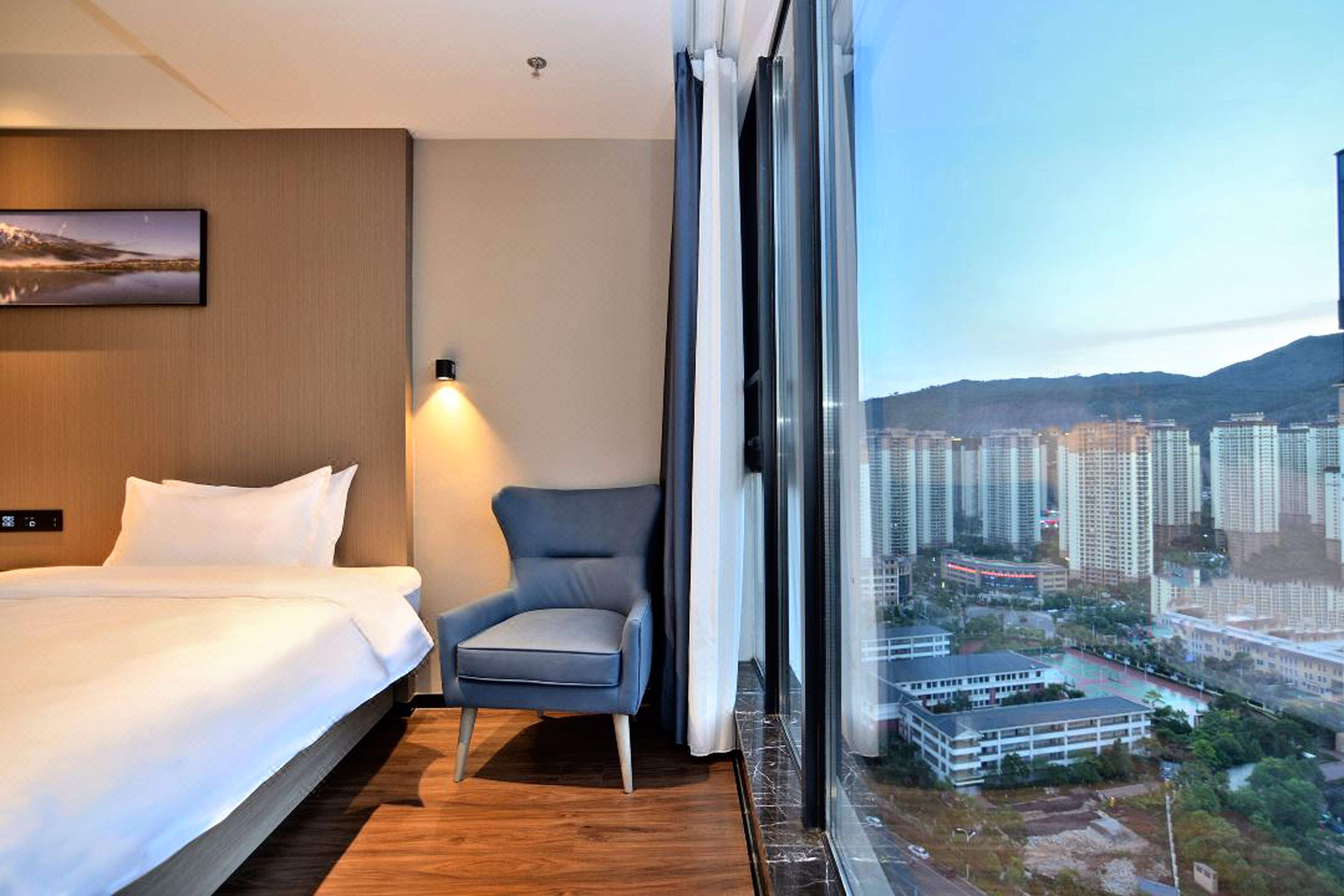 A hotel room with a bed, a chair, and a large window offering a city view.