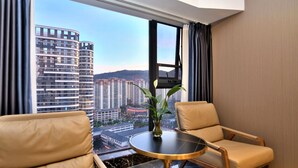 A hotel room with a view of tall buildings, a round table, two chairs, and a vase with a plant. - kunningyuehaojiudian (Kunming)