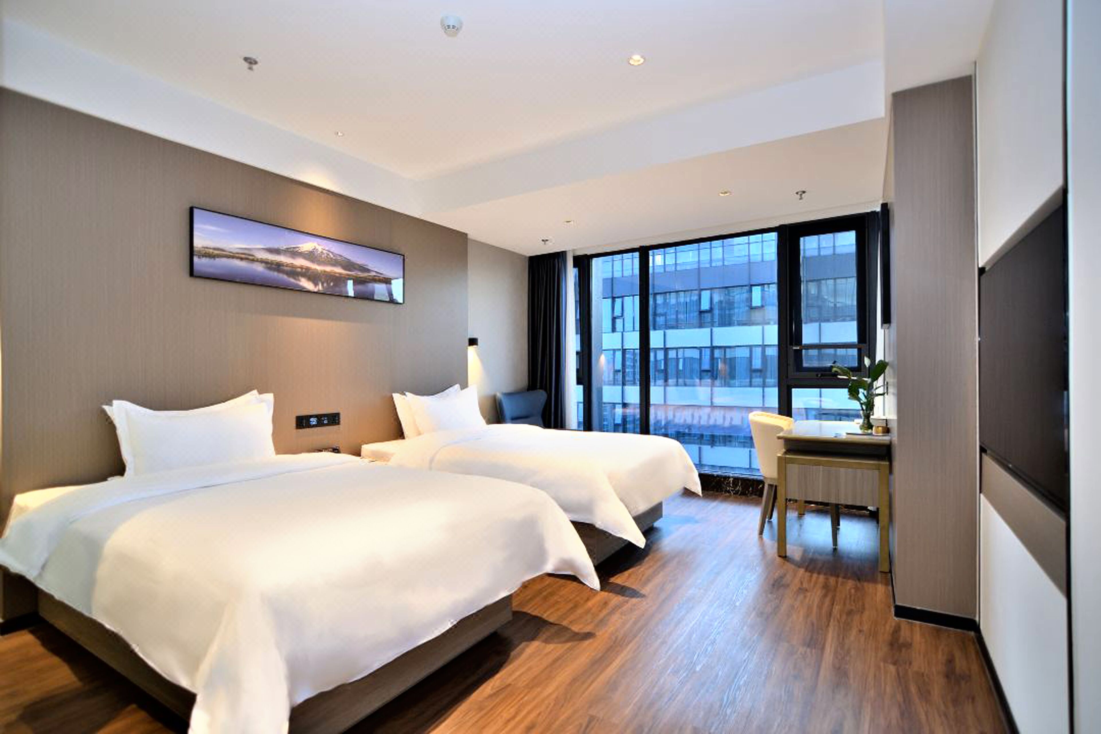 A modern hotel room with two beds, a desk, and a large window offering a city view.