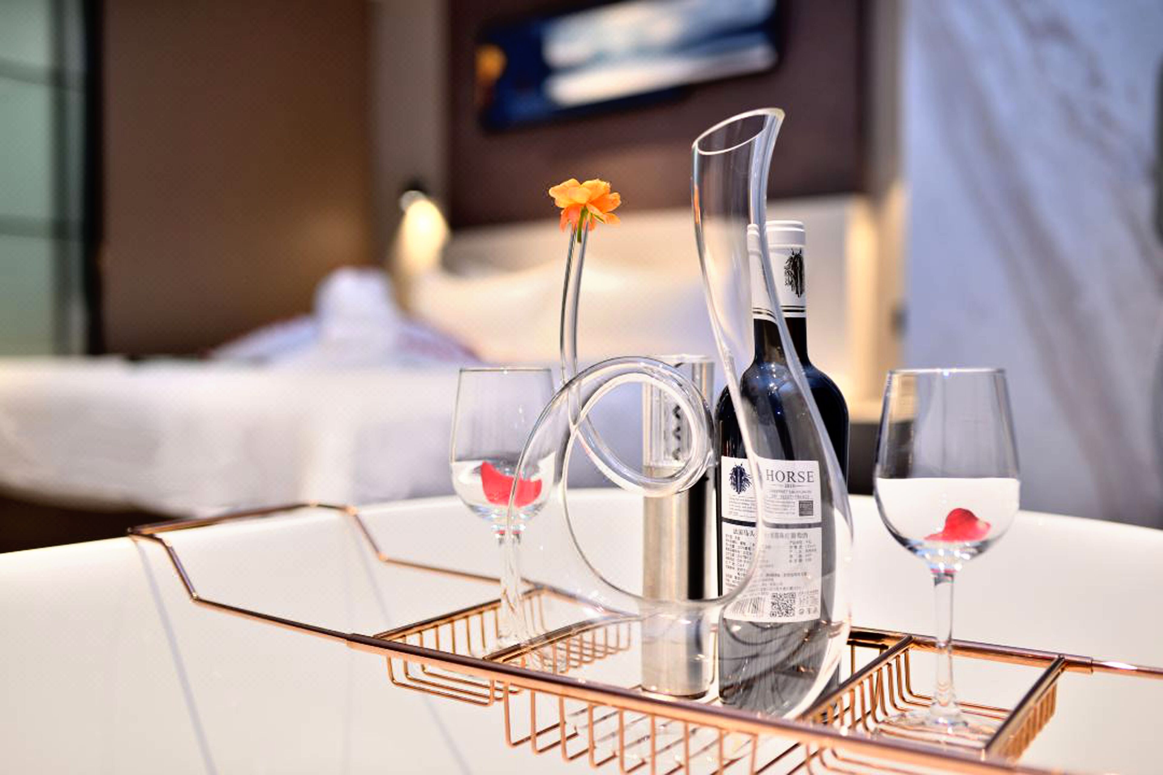 A hotel room with a bed, a bedside table with a bottle and glasses, and a flower on the table.