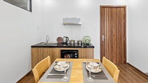 A compact kitchen with a black countertop, wooden cabinets, and a built-in dishwasher. There is a dining table with two chairs and a small window above it. - TCM Hotel & Apartment (Da Nang)