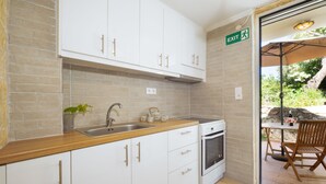 A compact kitchen with white cabinets, a wooden countertop, a sink, and an oven. There's a green exit sign on the wall and a patio area with a table and chairs visible through an open door. - Galatas Maisonette (Galatas)