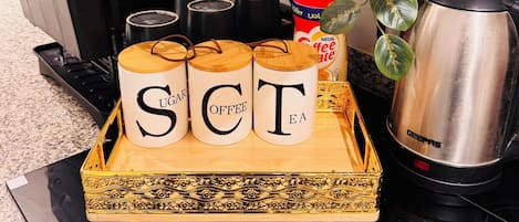 A kitchen counter with a tray of labeled containers for sugar, coffee, and tea, a coffee machine, a kettle, and a plant.