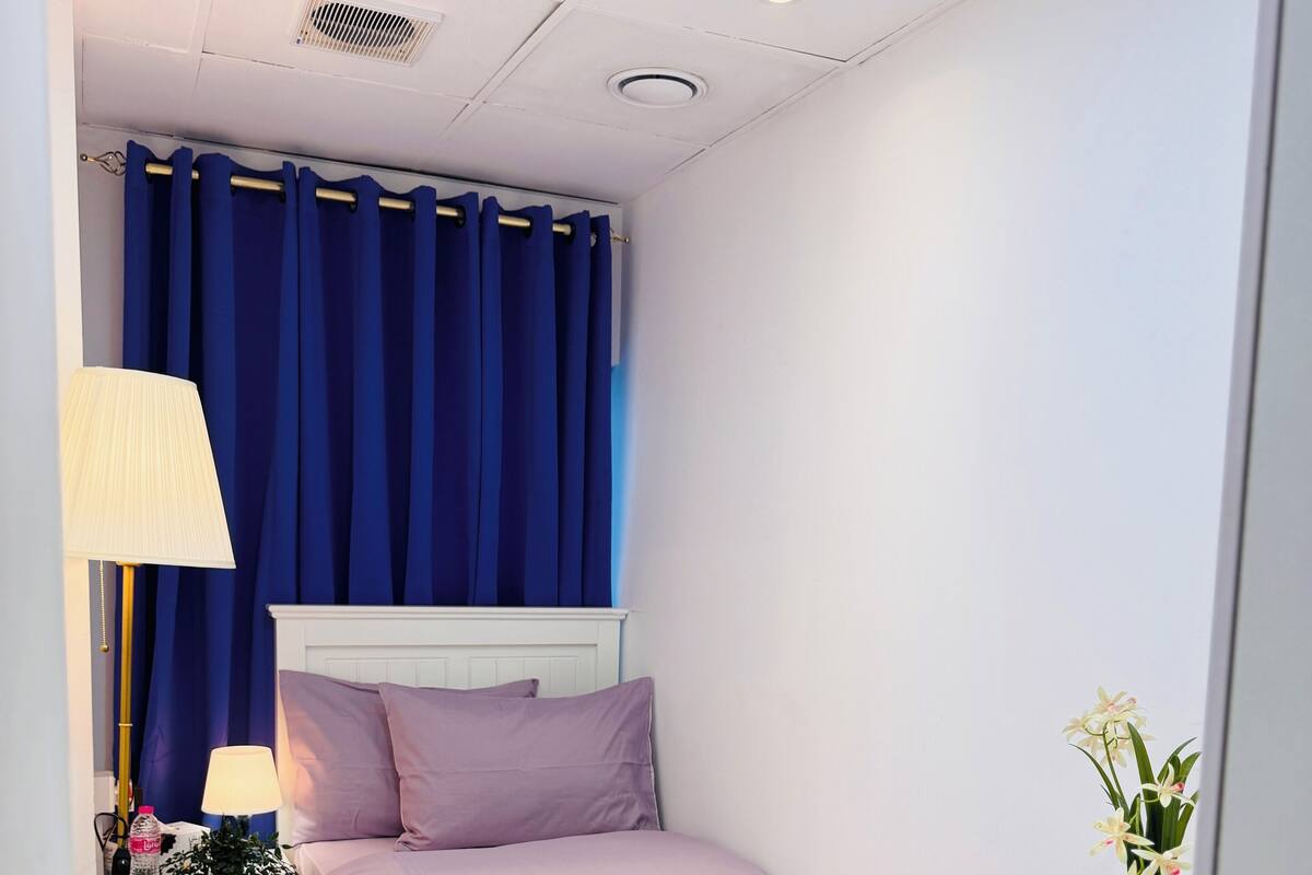 A bedroom with a bed, bedside table, chair, lamp, curtains, and a potted plant.