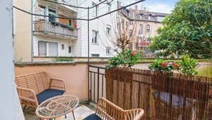 A balcony with wicker furniture, a small table, and a view of neighboring buildings and a tree. - Central Karlsruhe Apt: Balcony, Fast WiFi (Karlsruhe)
