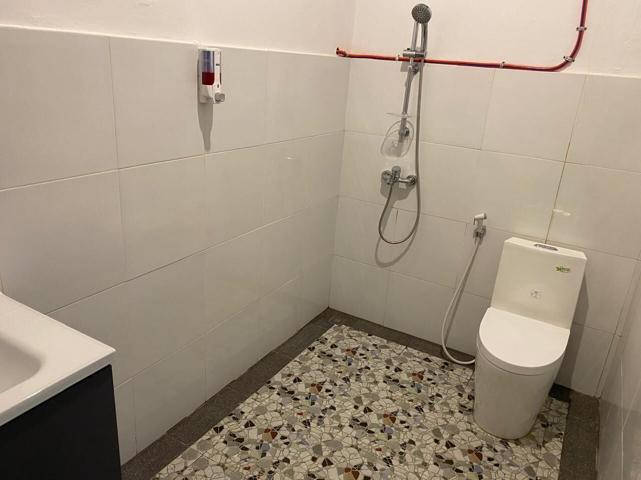 A small bathroom with white tiled walls, a mosaic floor, a toilet, and a shower with a red pipe.