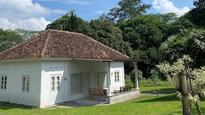 A white building with a tiled roof, a porch with wooden furniture, and a grassy lawn. - BADAI HERITAGE VILLA KALIURANG (Pakem)
