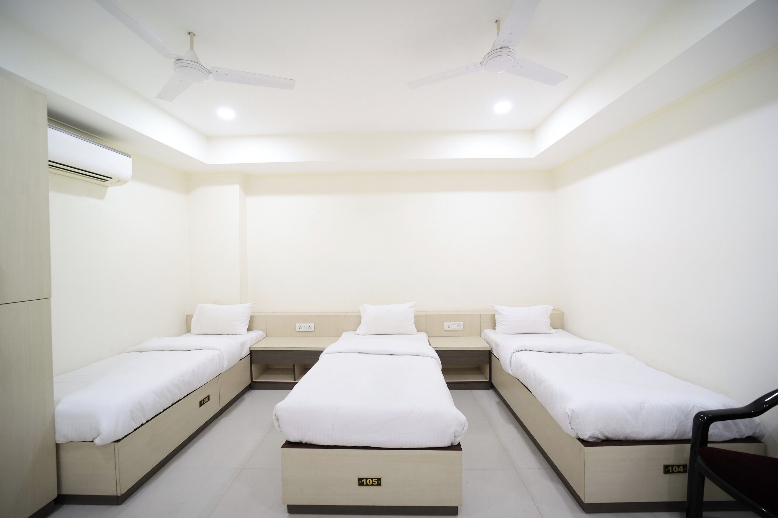 Une chambre d’hôtel avec trois lits simples, un ventilateur de plafond, un appareil de climatisation et une chaise.