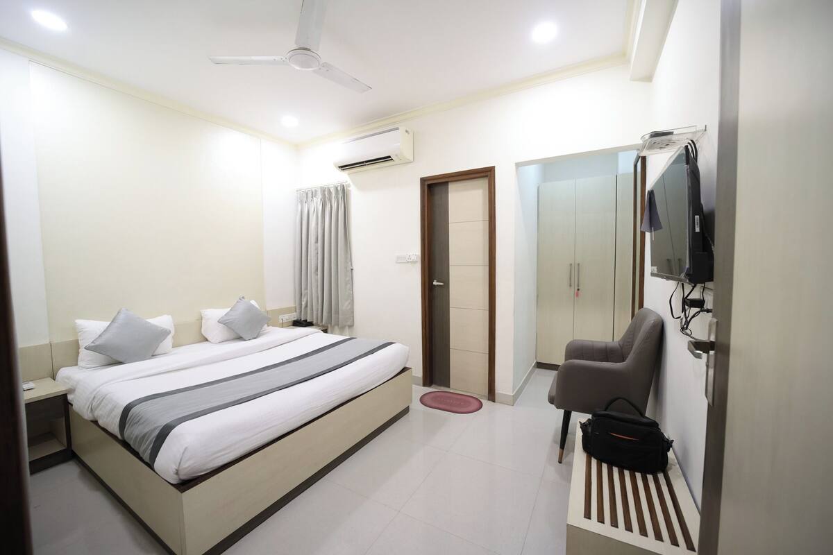 A hotel room with a bed, a chair, a TV, an air conditioning unit, a ceiling fan, and a door.