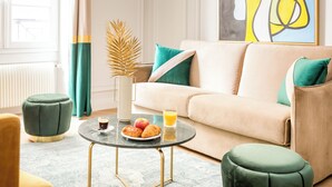 Un salon avec un canapé beige, des coussins verts, une table basse ronde sur laquelle se trouve une assiette de croissants et un verre de jus d’orange, ainsi qu’un vase décoratif en forme de feuille de palmier.