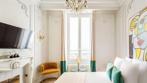 Une chambre d’hôtel avec un grand lit, une télévision, une petite table avec un vase, un fauteuil jaune et une vue sur le balcon.