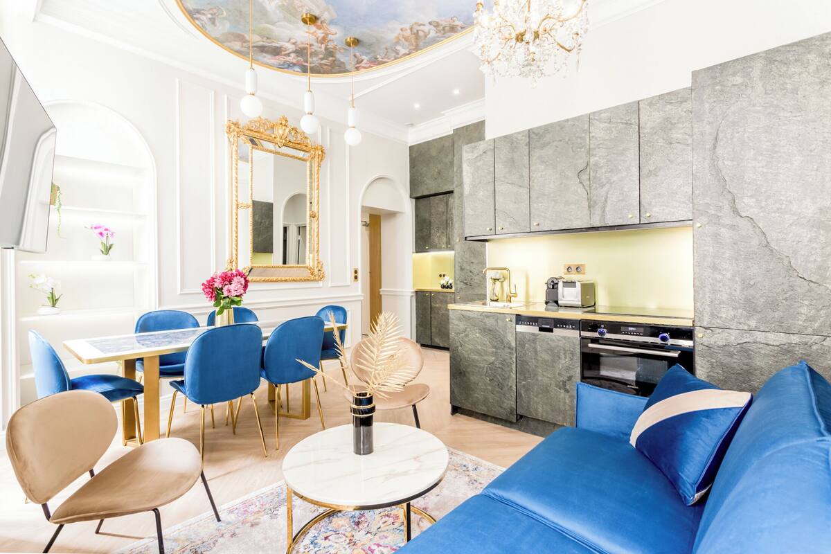 A modern living room with a blue sofa, a dining table with chairs, a large mirror, and a kitchen area with a built-in oven.