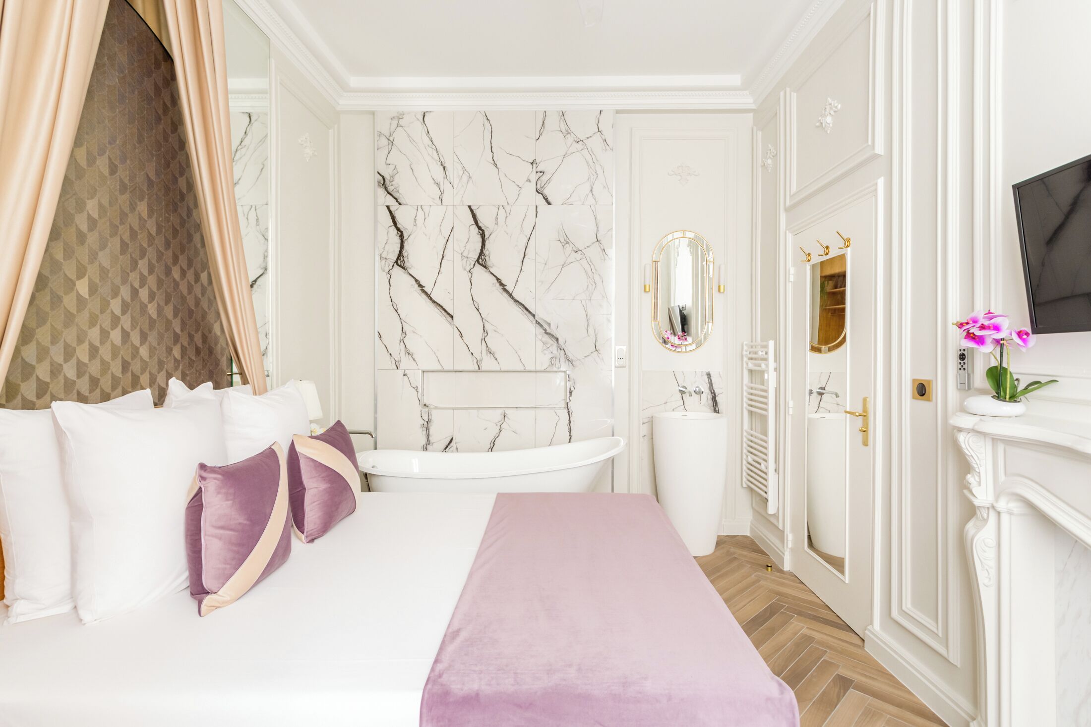 A luxurious bedroom with a large bed, a marble wall, a white bathtub, and a TV mounted on the wall.