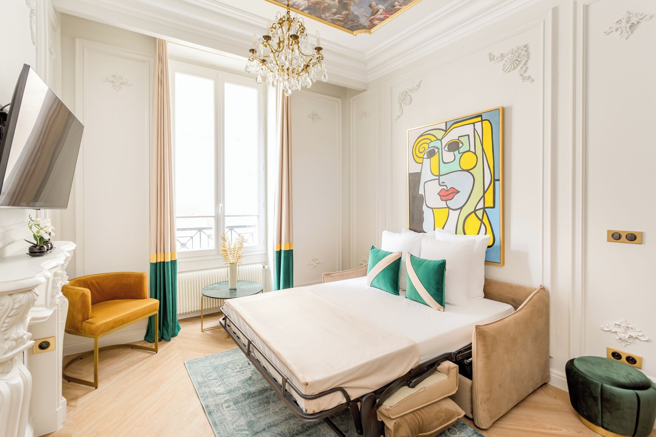 A hotel room with a bed, a TV, a chair, a painting, and a view of the city.