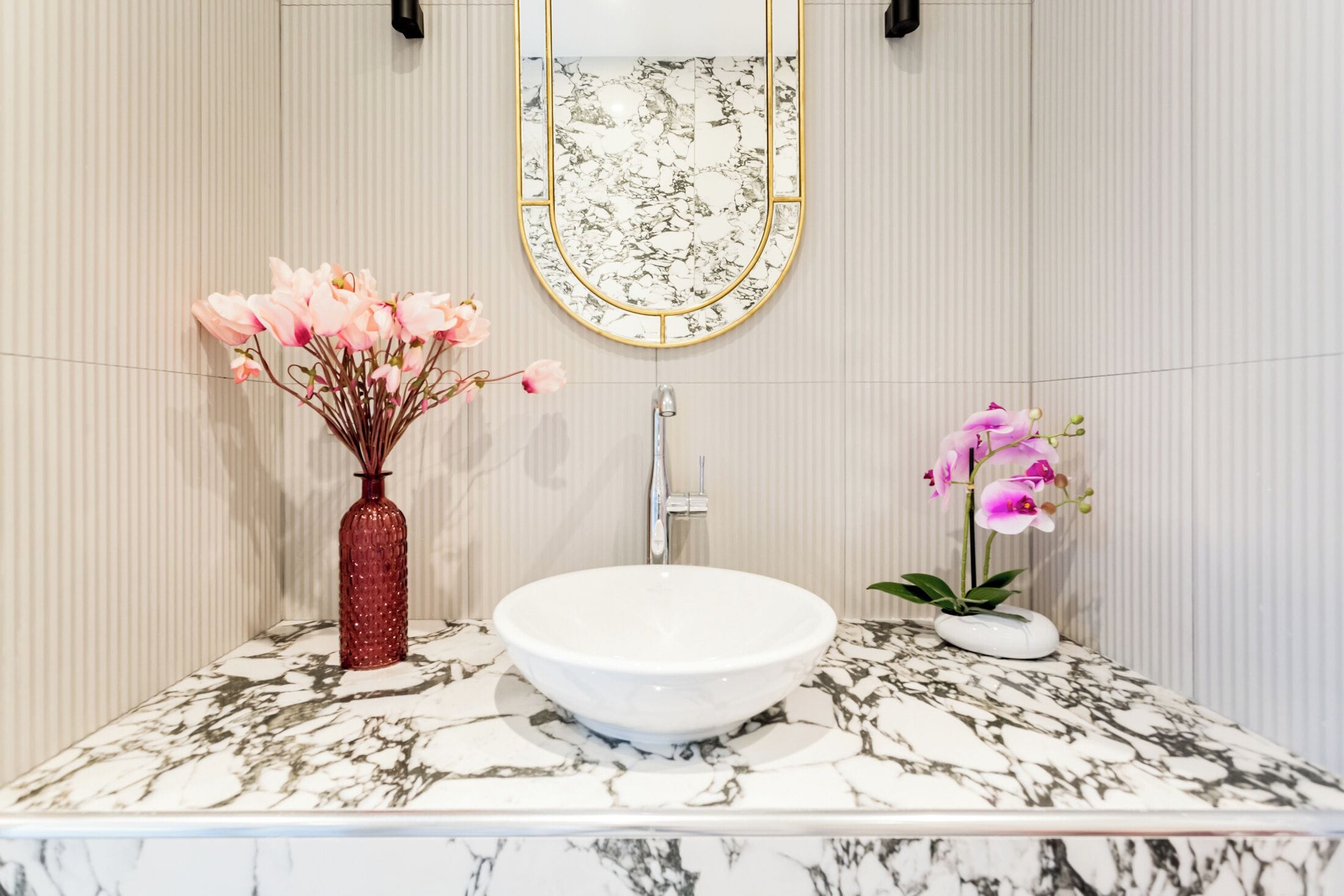 Une salle de bain avec un lavabo en marbre, un vase rouge contenant des fleurs roses et un petit pot blanc avec une orchidée rose.