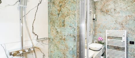 A modern bathroom with a marble wall, a shower with a handheld showerhead, a white sink, and a towel rack.