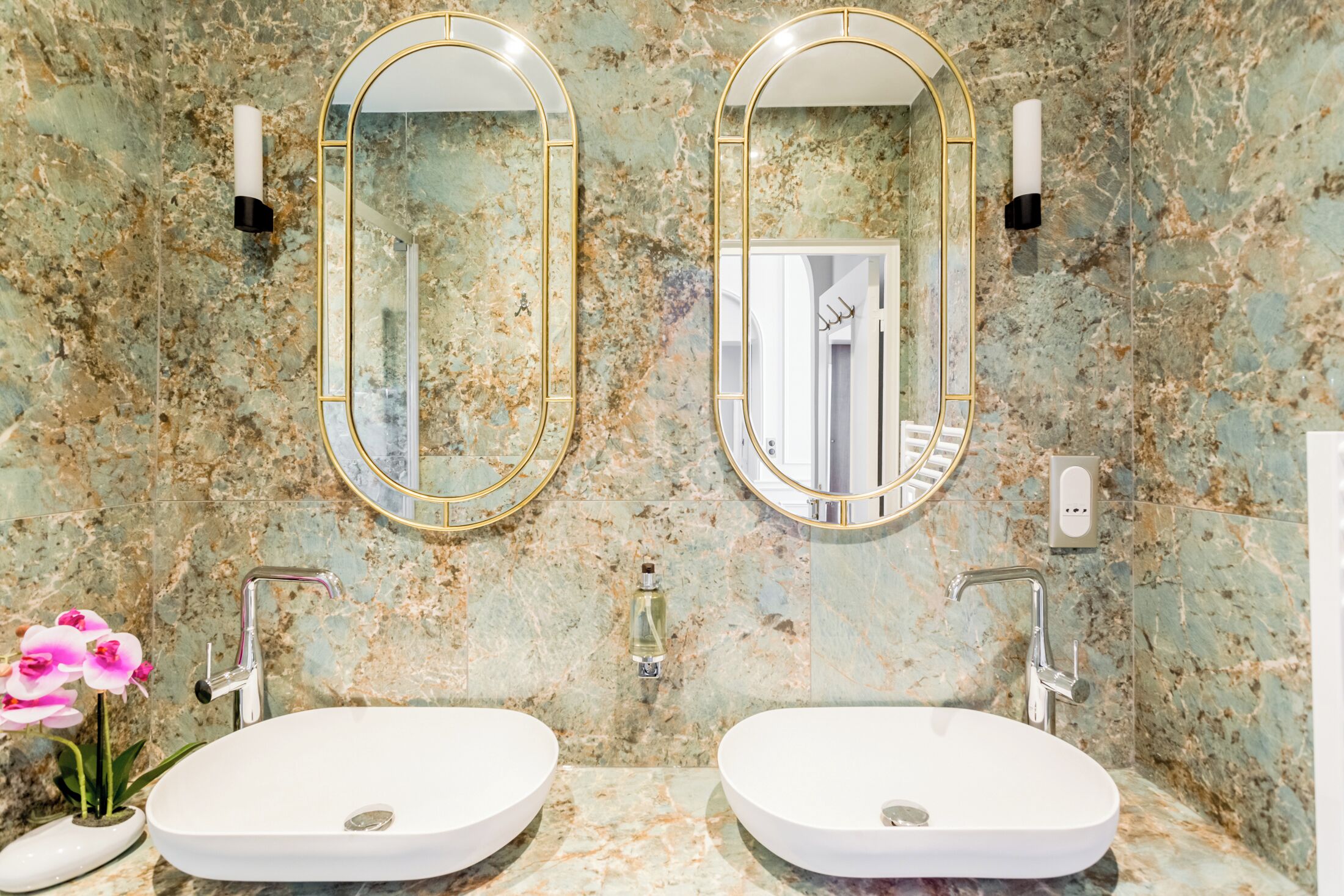 A bathroom with two oval sinks, marble walls, and gold-framed mirrors.