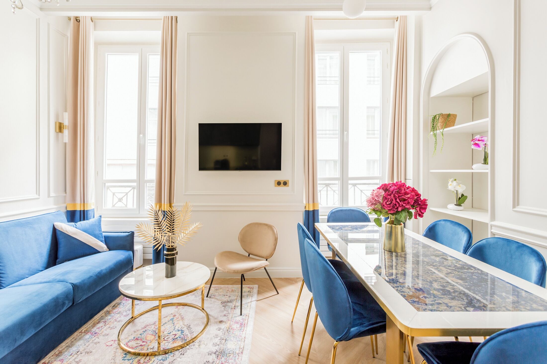 A modern living room with a blue sofa, a round coffee table, a dining table with blue chairs, and a TV mounted on the wall.