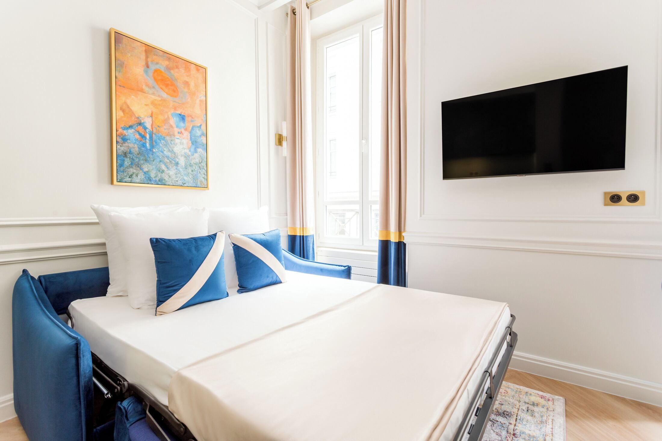 A hotel room with a bed, a flat-screen TV, a painting, and a window with curtains.