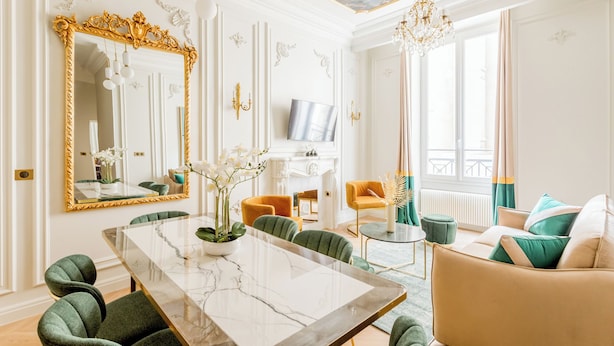 Un vaste salon comprenant une table à manger en marbre, des chaises recouvertes d’un tissu vert, un canapé beige et un téléviseur fixé au mur.