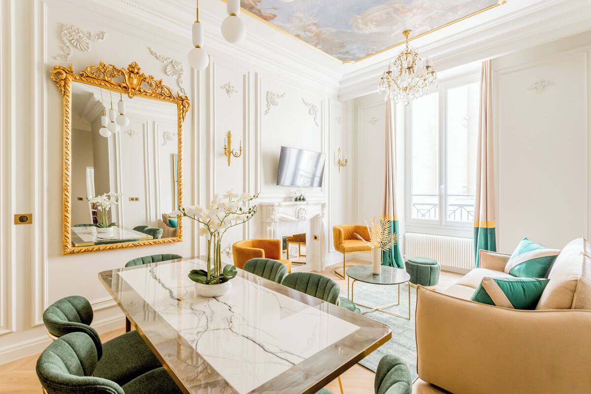 A spacious living room with a marble dining table, green upholstered chairs, a beige sofa, and a television mounted on the wall.
