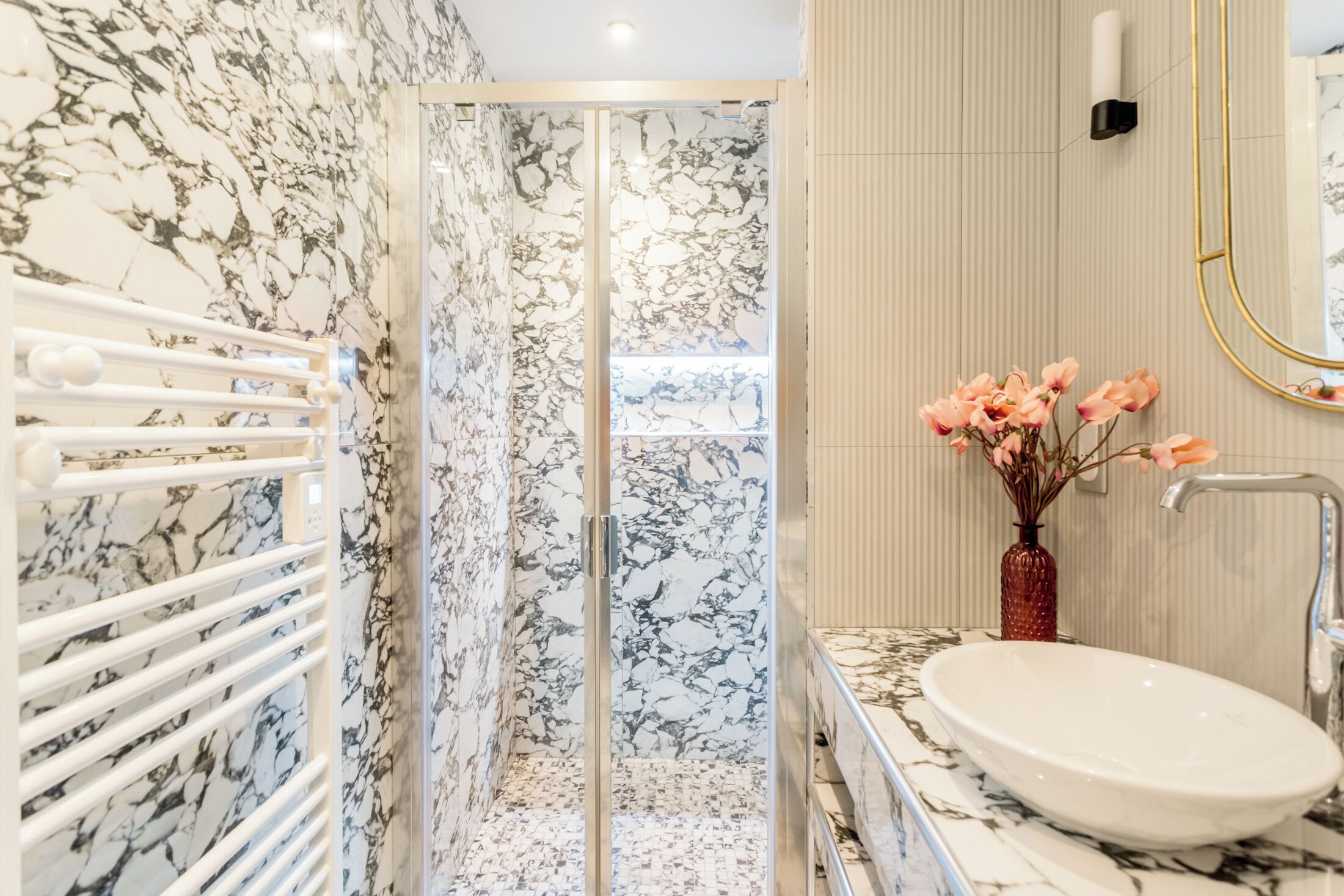 Une salle de bain avec un mur en marbre, une cabine de douche en verre, un lavabo blanc et un vase rempli de fleurs roses.