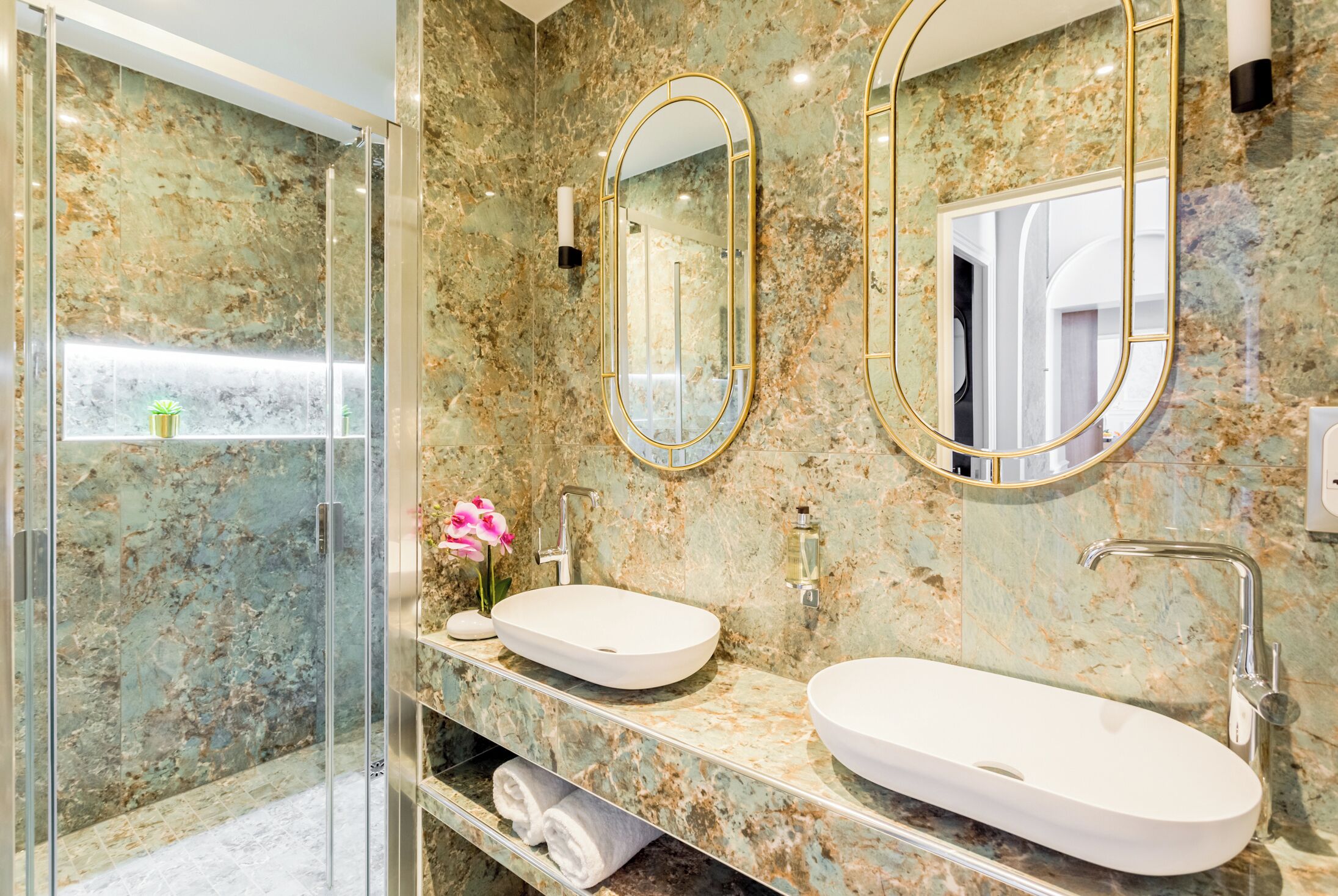 A modern bathroom with a glass-enclosed shower, a white oval sink, and two mirrors with gold frames.