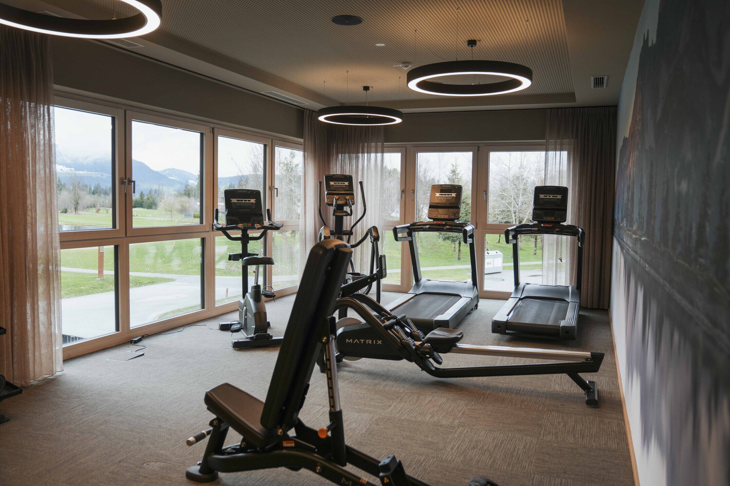 Un gimnasio con cintas de correr, una máquina de remo y equipo de pesas. Amplias ventanas ofrecen vistas a un parque y montañas.