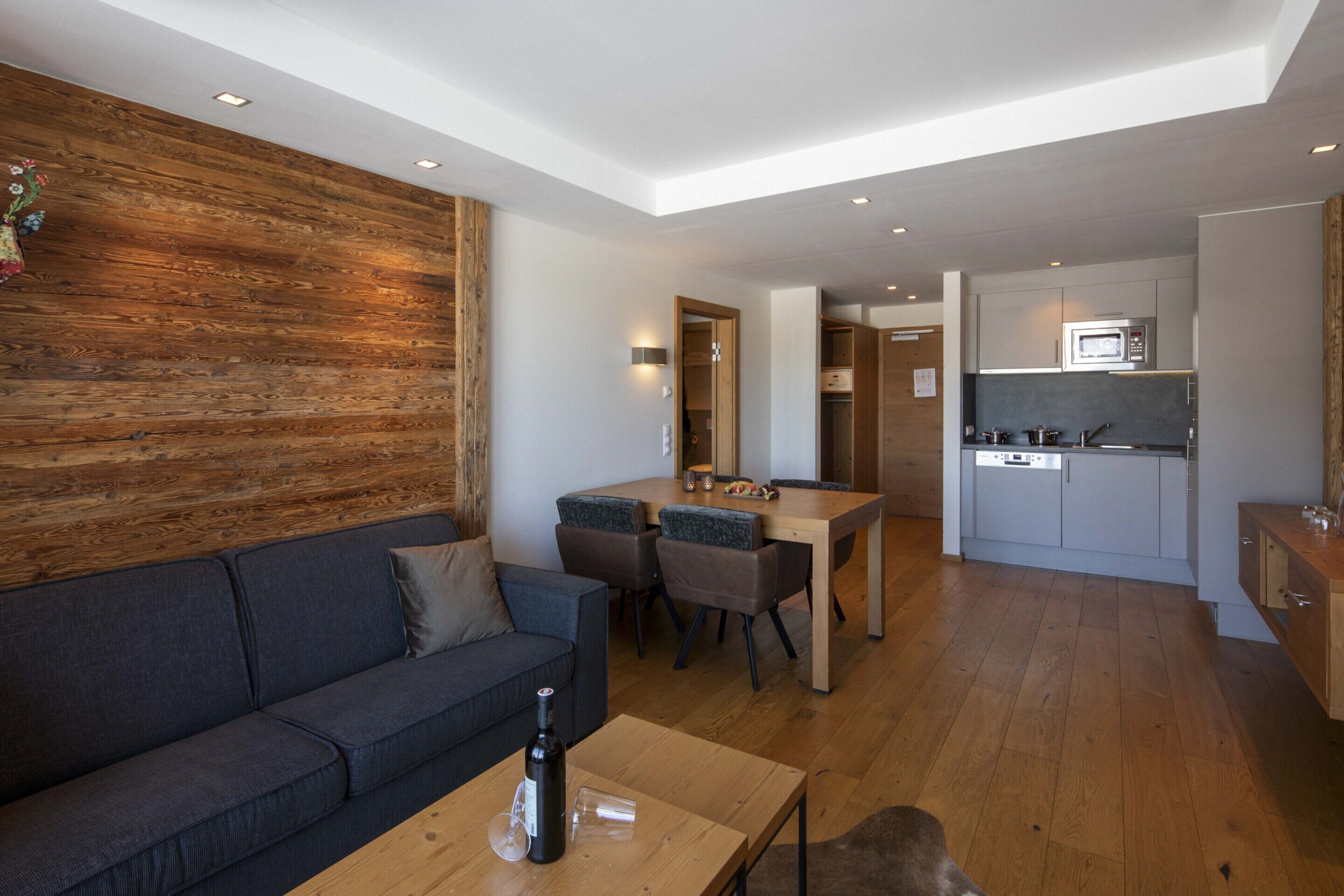 A modern living room with a sofa, a wooden accent wall, a dining area with chairs, and a kitchen with cabinets and a microwave.