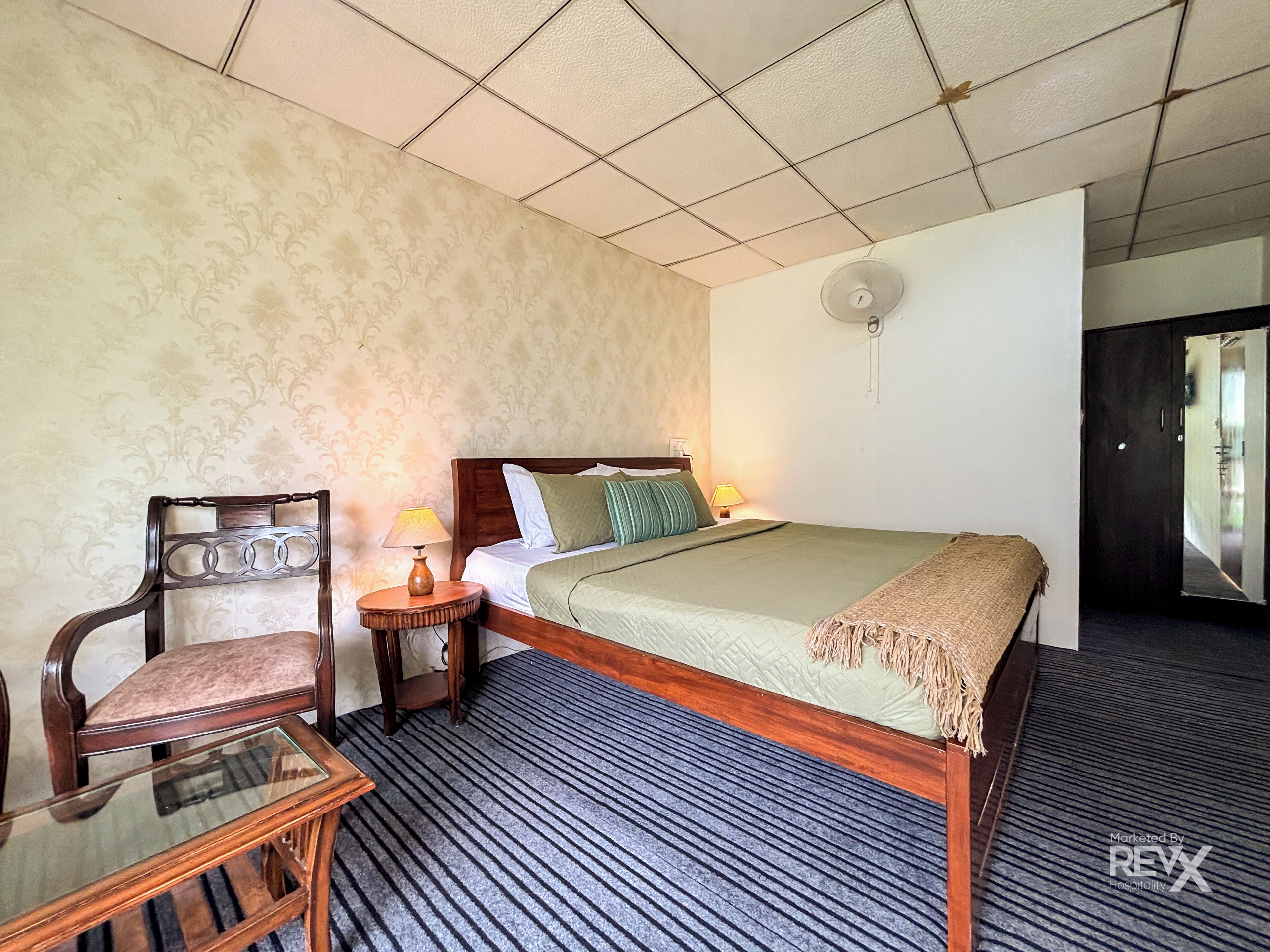 A hotel room with a bed, a chair, a small table, a lamp, and a ceiling fan.