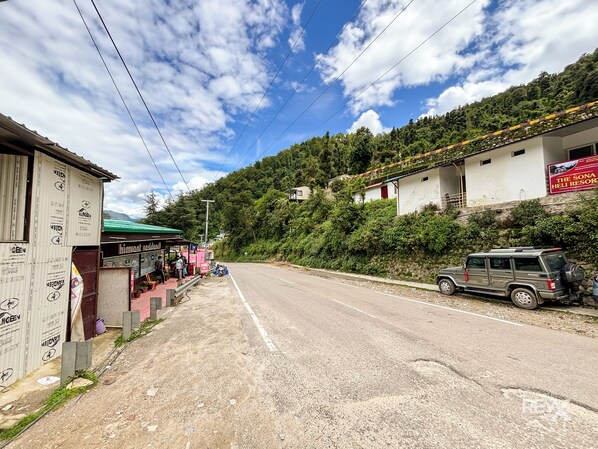 Una carretera con una camioneta SUV estacionada, una pequeña tienda a un lado del camino y un edificio con un letrero que dice 'The Sona Helia Resort'.