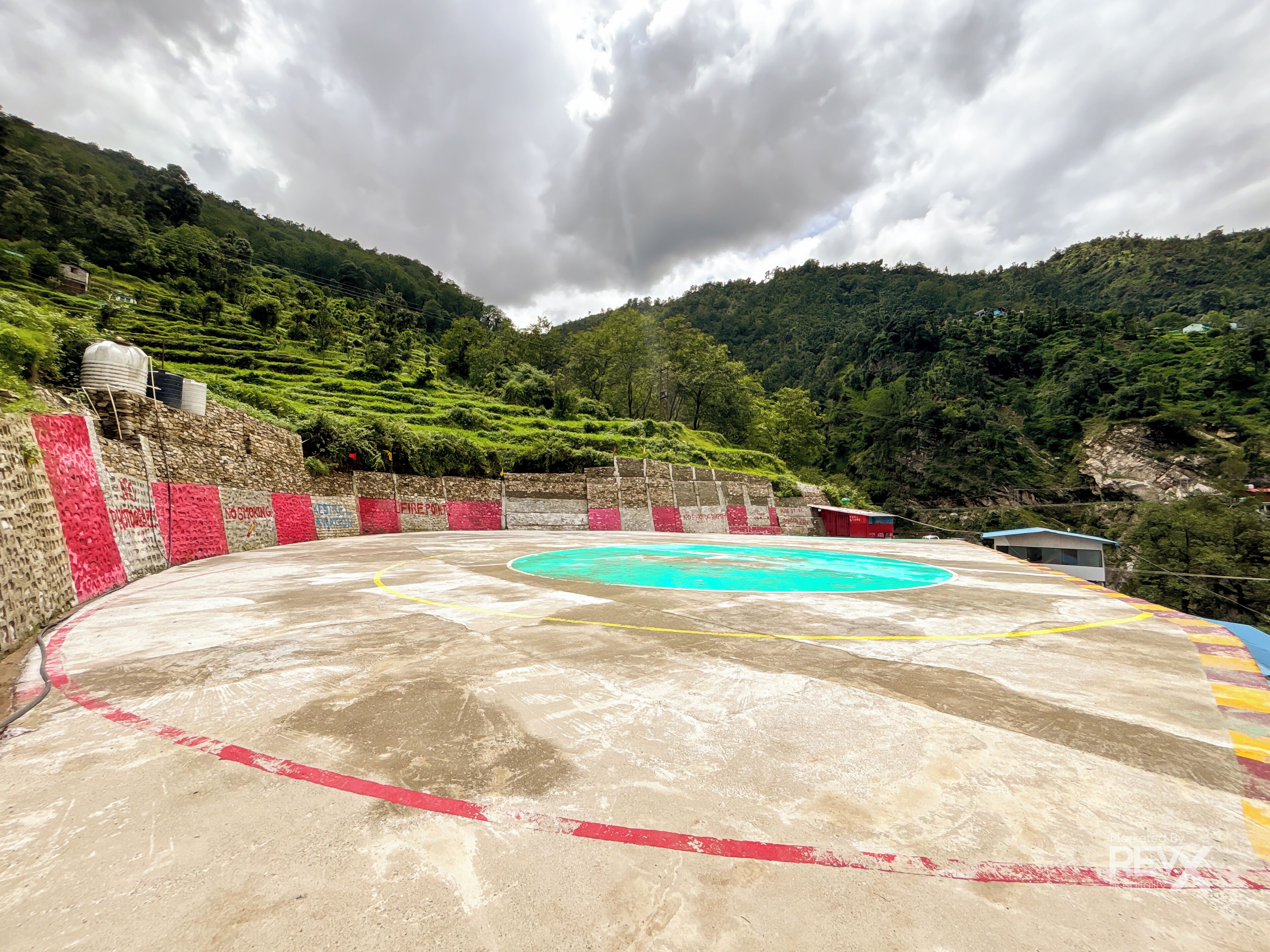 A concrete sports court with red and yellow markings, surrounded by a stone wall with graffiti, set against a backdrop of lush green hills and a cloudy sky.