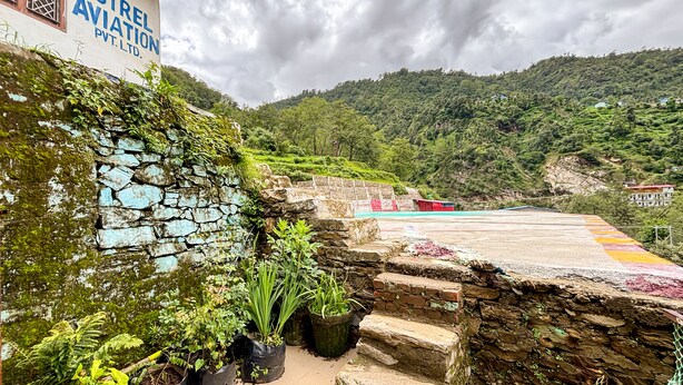 Un muro de piedra con vegetación, un edificio con la señalización 'Kestrel Aviation Pvt. Ltd.' y un paisaje montañoso al fondo.