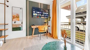 En moderne stue med et fladskærms-tv monteret på væggen, et træskrivebord med stol, et sofabord i glas med en vase, og et stort vindue med gardiner.