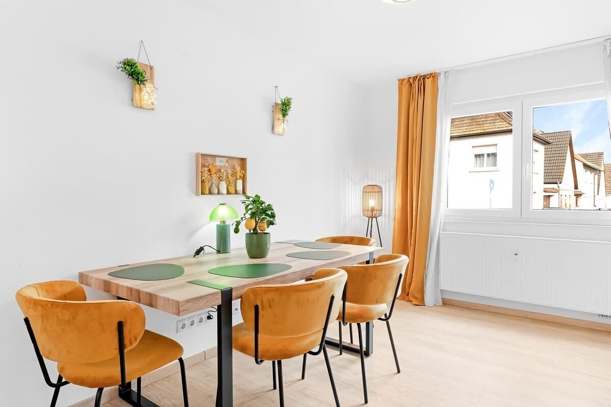 A dining area with a wooden table, six chairs, and a view of the outside through a window with curtains.