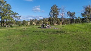 A small, single-story cabin with a sloped roof, surrounded by a grassy field and trees. - Bickell's Hut - Off-Grid Farm Stay, Fire Pit, Fire Bath, Cozy, Views - Sleeps 2 -4 (Hydes Creek)