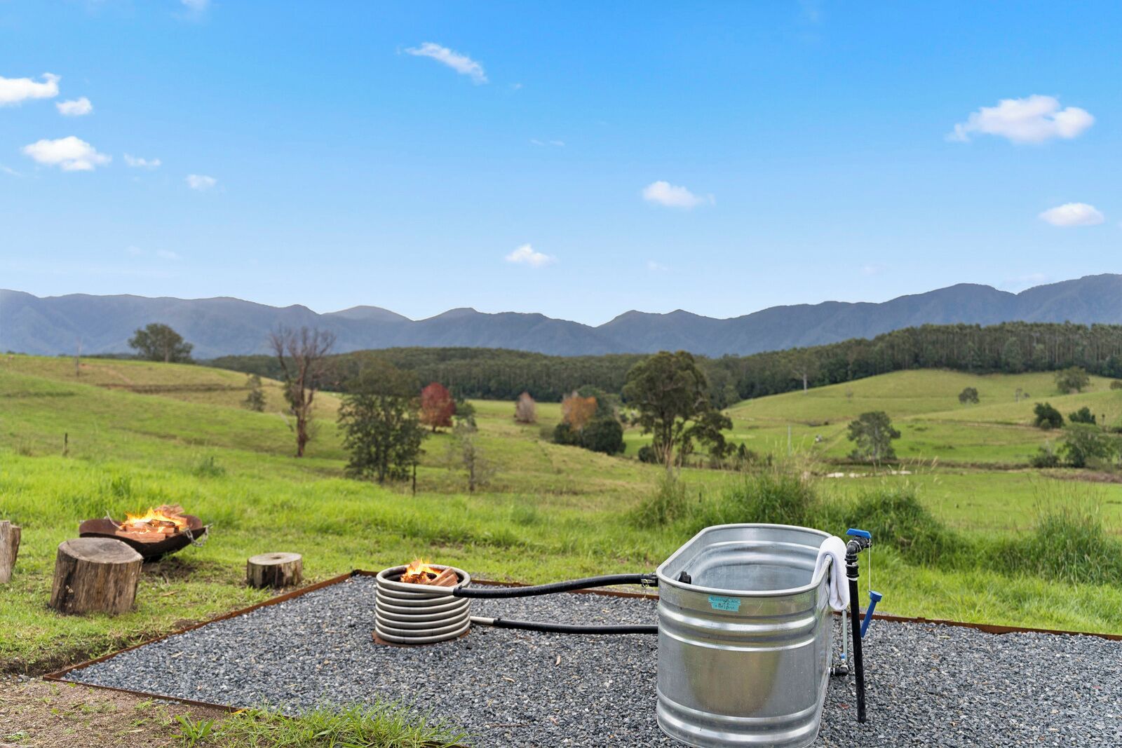 A campsite with a fire pit, a metal barrel, and a hose, set against a backdrop of green hills and mountains.