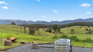 A campsite with a fire pit, a metal barrel, and a hose, set against a backdrop of green hills and mountains.