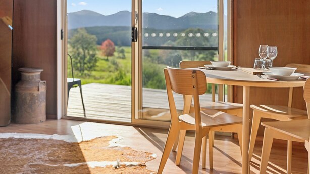 A dining area with a table set for two, wooden chairs, and a view of a mountainous landscape through large sliding glass doors. - Bickell's Hut - Off-Grid Farm Stay, Fire Pit, Fire Bath, Cozy, Views - Sleeps 2 -4 (Hydes Creek)