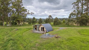 A small, single-story house with a curved roof, situated in a grassy field with trees in the background. - Bickell's Hut - Off-Grid Farm Stay, Fire Pit, Fire Bath, Cozy, Views - Sleeps 2 -4 (Hydes Creek)