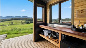 A wooden cabin with a kitchen area, large windows, and a view of a green landscape. - Bickell's Hut - Off-Grid Farm Stay, Fire Pit, Fire Bath, Cozy, Views - Sleeps 2 -4 (Hydes Creek)