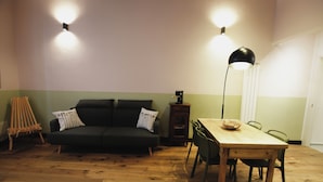 Ein Zimmer mit Holzboden, einem dunkelgrünen Sofa mit Kissen, einer kleinen Holzkabine, einem Esstisch mit Stühlen und zwei Wandleuchten.