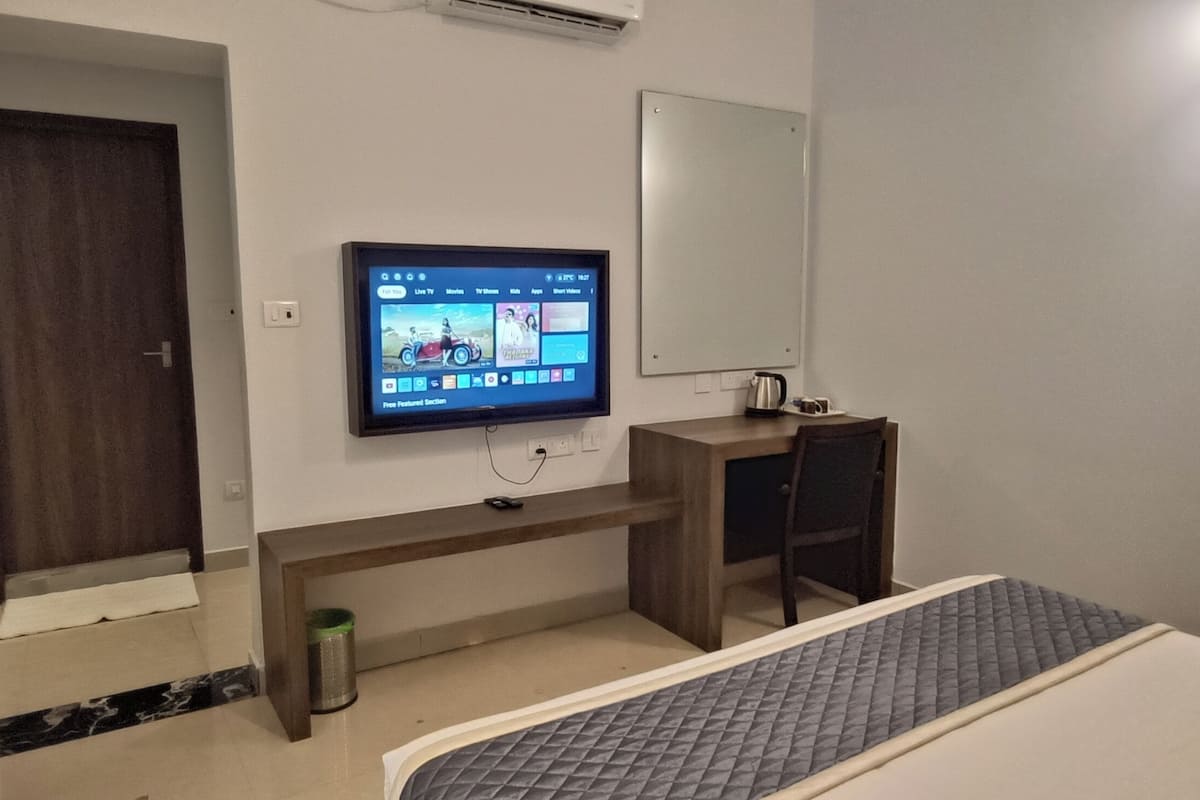 A hotel room with a TV mounted on the wall, a desk with a chair, a small trash bin, and a bed with a mattress.