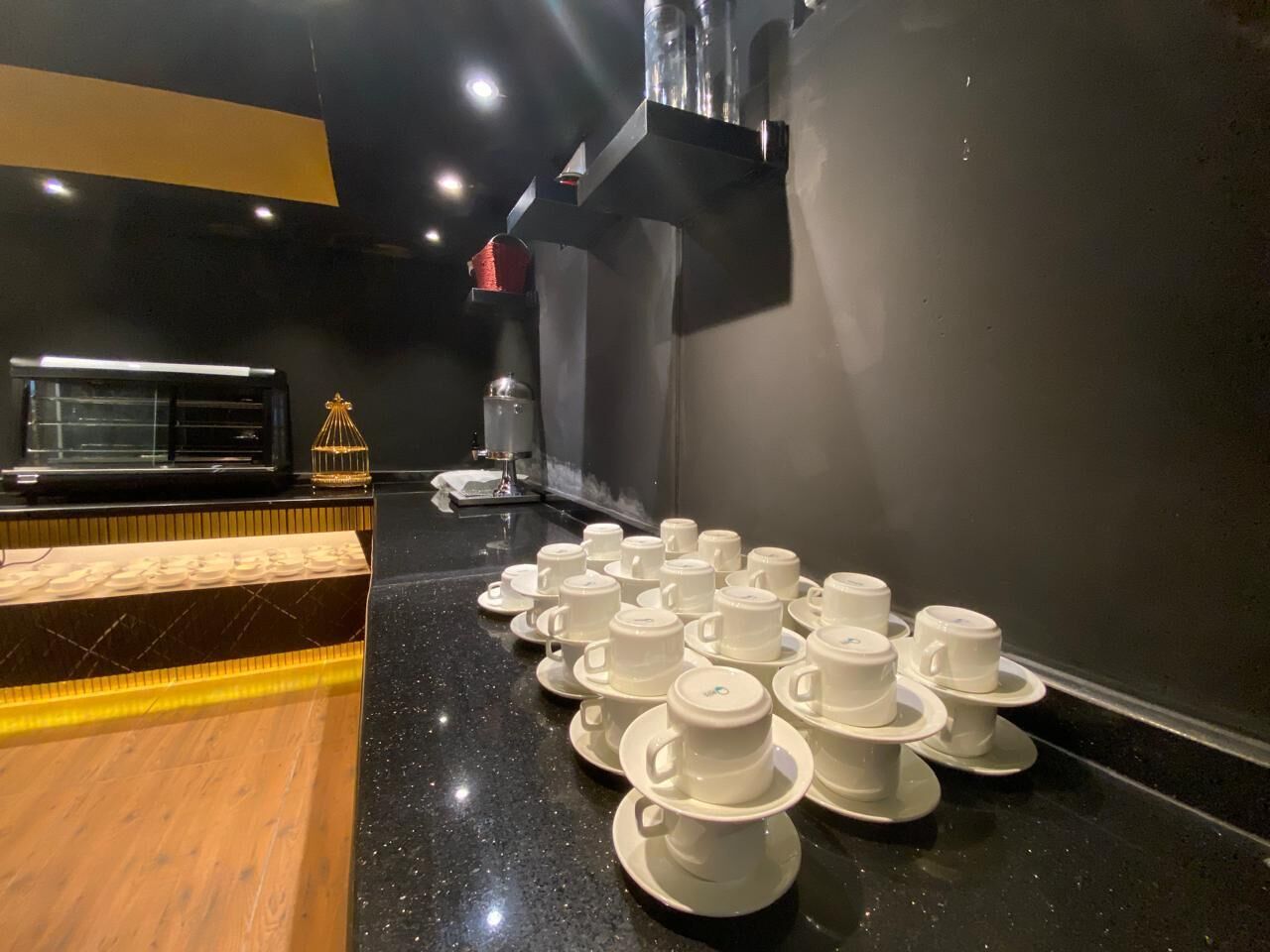 A room with a black countertop, white cups and saucers arranged on it, and a coffee machine in the background.
