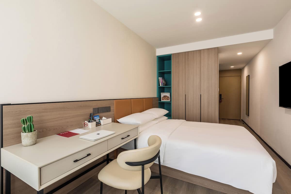 Habitación de hotel con cama, escritorio con silla, una pequeña planta en maceta y televisor montado en la pared.