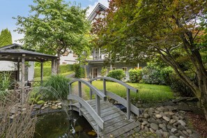 Un puente de madera sobre un estanque, rodeado de abundante vegetación y una casa con jardín.