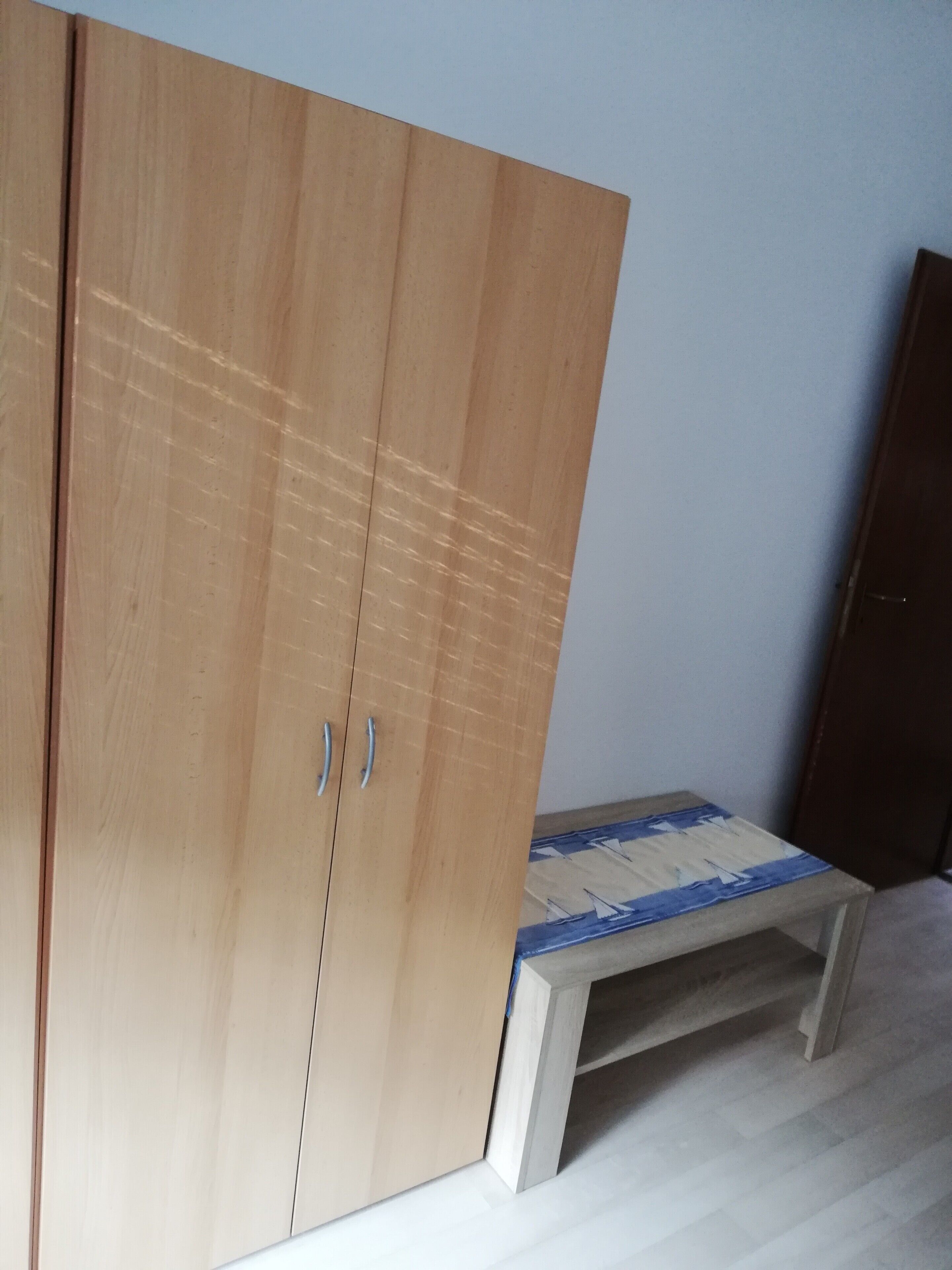 A room with a wooden wardrobe, a small table with a blue and white patterned tablecloth, and a brown door.