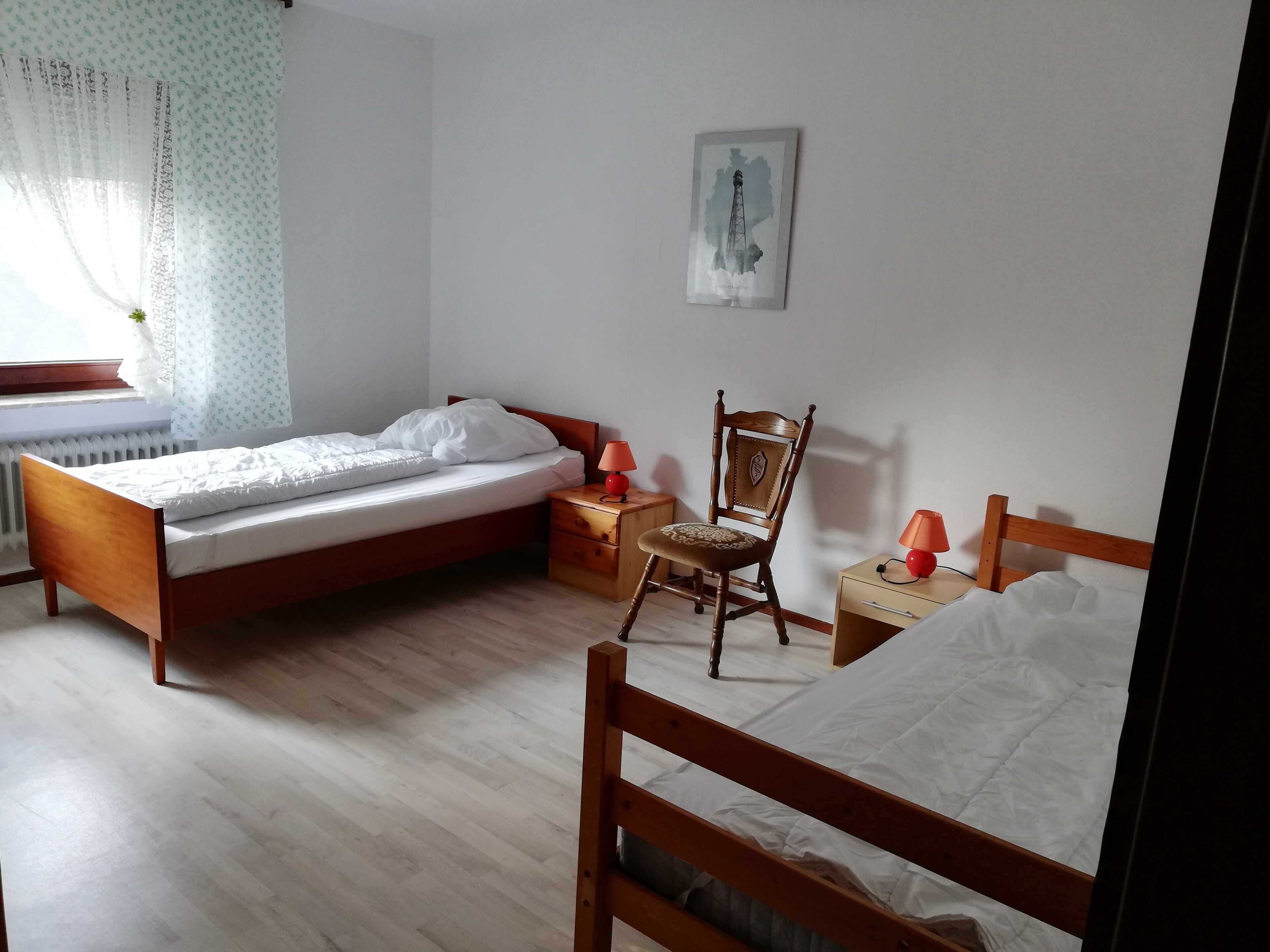 A bedroom with a wooden bed, a wooden chair, a bedside table with a lamp, and a window with curtains.
