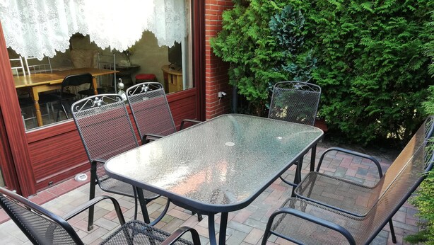En terrasse med et bord med glasplade og metalstole, en murstensvæg og en grøn busk.
