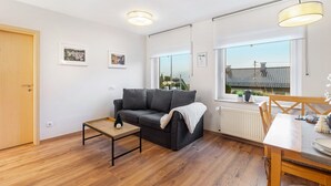 A modern living room with a sofa, a small table, a dining table, and a window with a view of buildings.